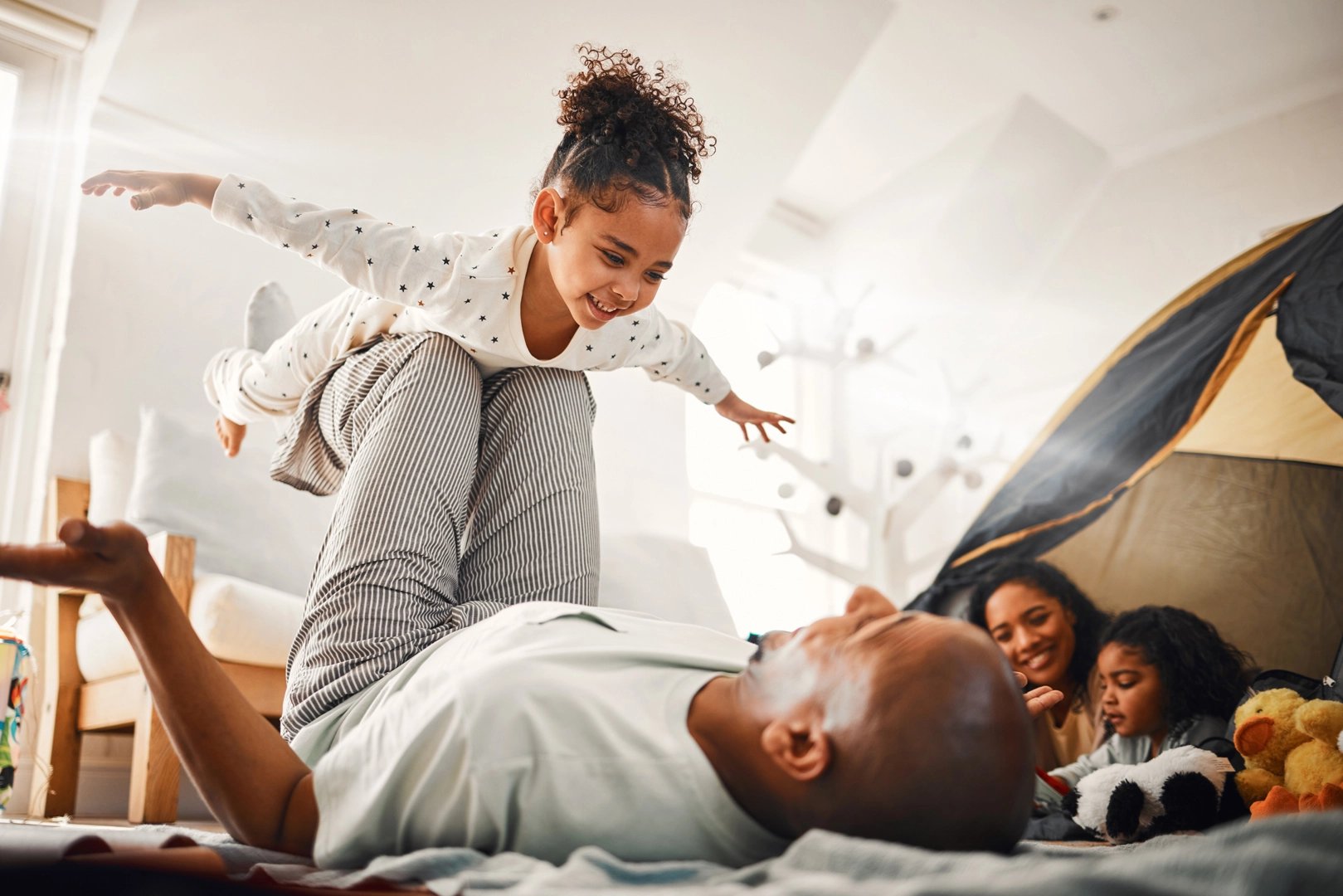 Père faisant faire l'avion à sa petite fille alors que la mère et le frère regardent
